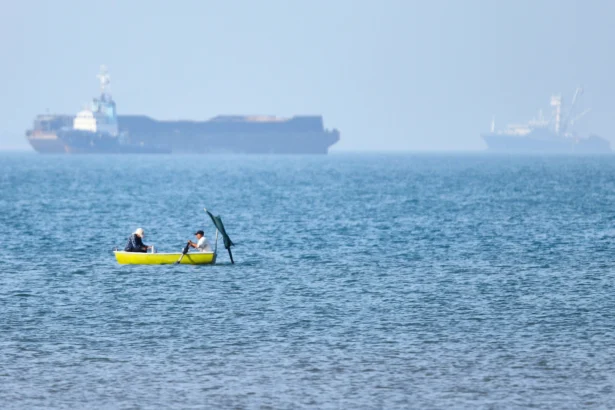Two men sit in a small boat on the water as cargo ships are anchored in the background in the Strait of Hormuz off Bandar Abbas, Iran, on May 4, 2026. (Amirhosein Khorgooi/ISNA via AP)